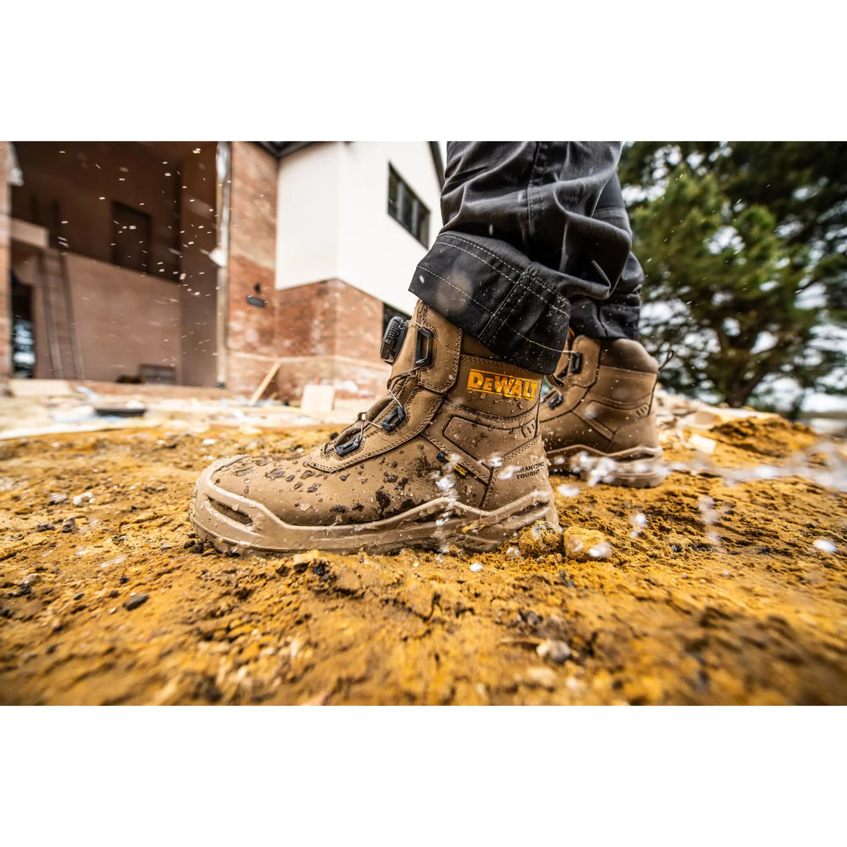 A close-up of a person wearing a pair of muddy DEWALT work boots standing on wet, splashing dirt outdoors